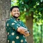 Portrait of a smiling man in a casual shirt leaning against a tree outdoors.