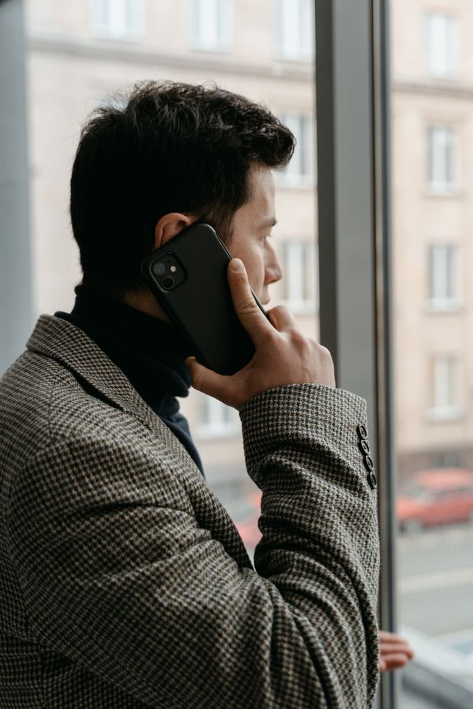Man in a checkered blazer using a smartphone while gazing out a window.